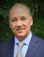 A professional headshot of a middle-aged man with a bald head, wearing a navy blue suit, light blue shirt, and patterned tie. He is smiling slightly against a soft-focus background of green foliage.