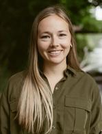 A smiling young woman with long blonde hair and a green button-down shirt stands in front of a blurred natural background. The outdoor lighting creates a warm, approachable feel for this professional headshot.