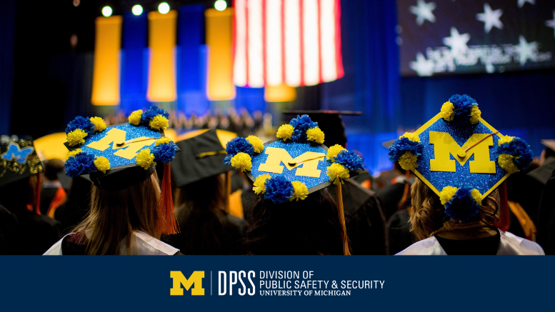 Graduates in caps decorated with blue and yellow flowers and block "M" face a stage at a University of Michigan ceremony; mood is celebratory.
